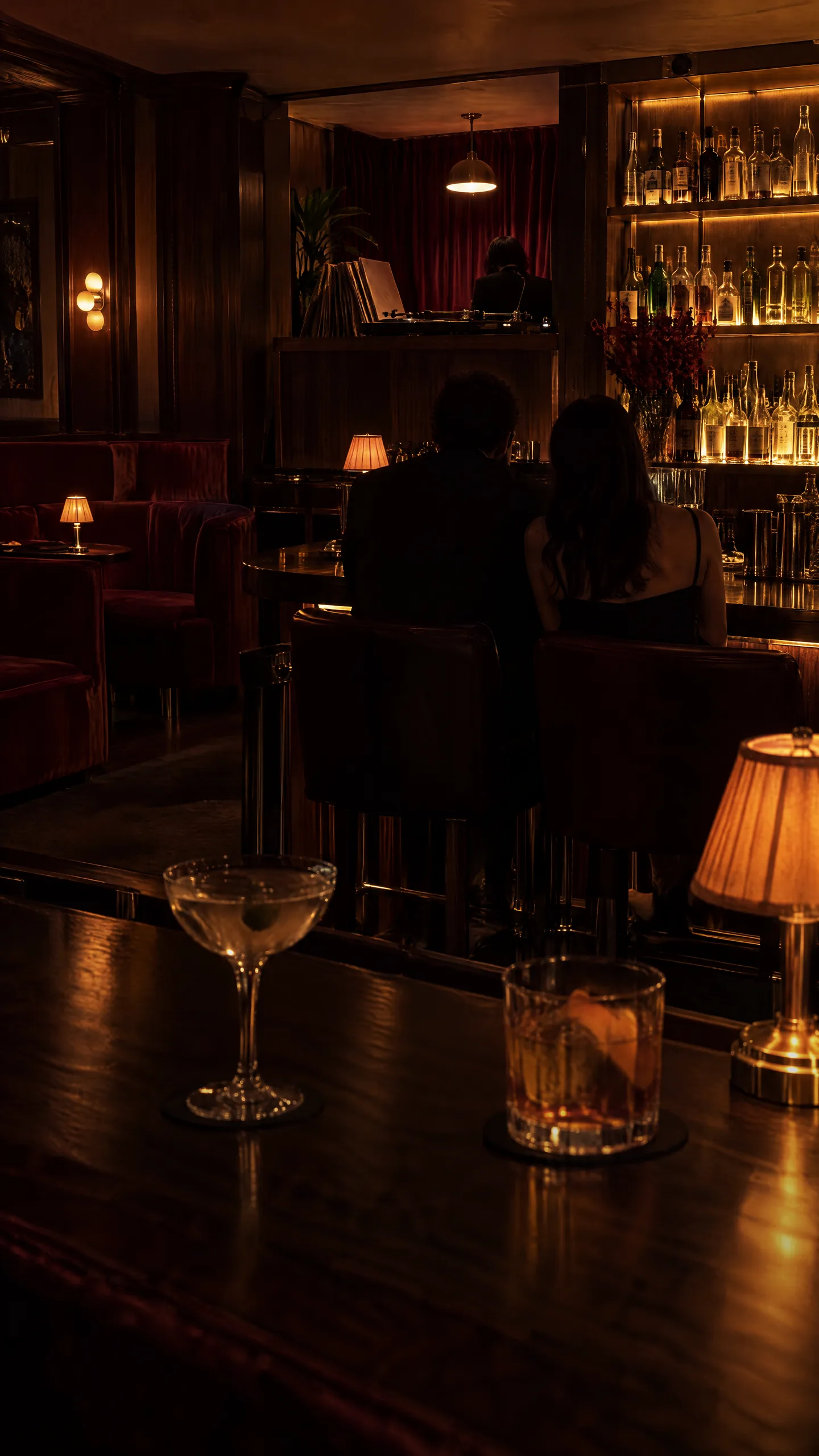 A low-lit cocktail bar: a couple at the counter, a drink and a lamp on the bar in front, a DJ playing records in the corner
