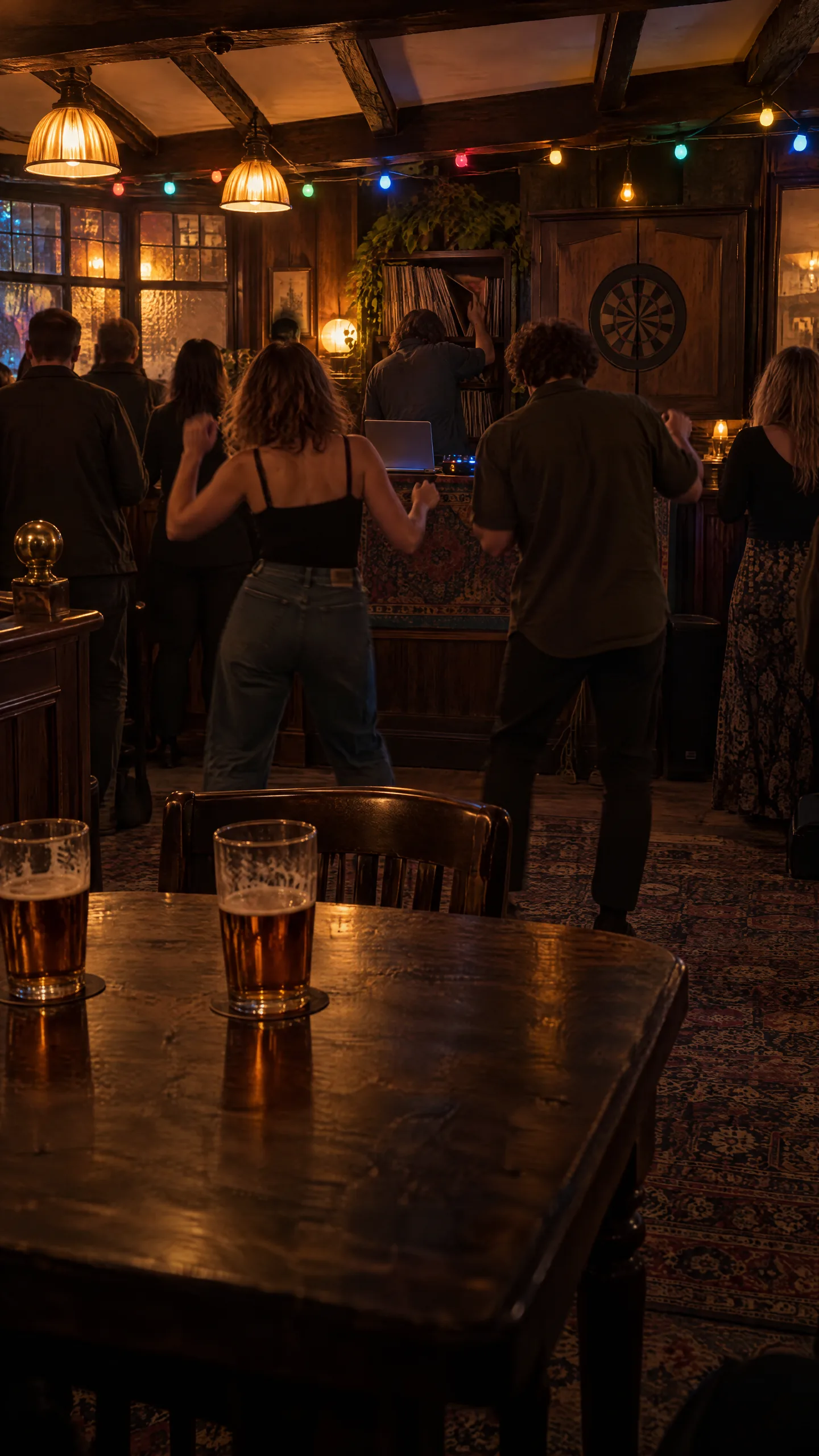 The back room of a pub: two pints on the table, people dancing on the carpet in front of the DJ, coloured bulbs and a dartboard